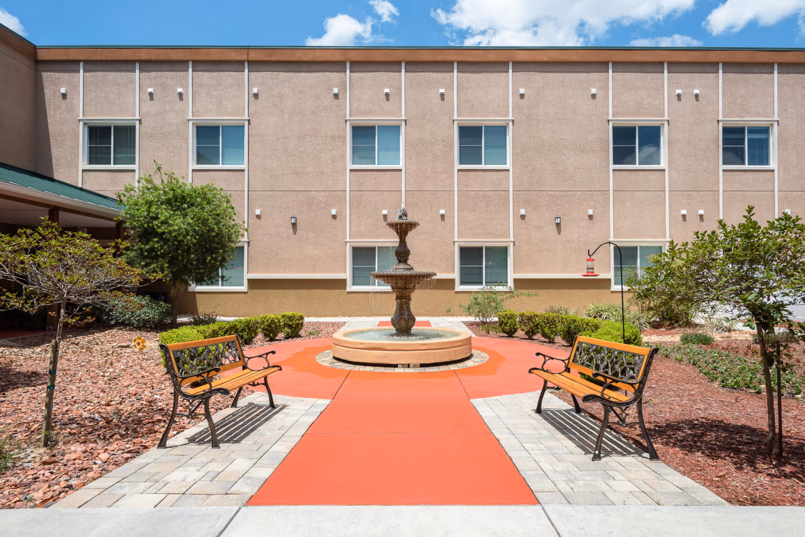 Outdoor courtyard area with a central water fountain surrounded by a red paved circular path. Two wooden benches with decorative metal armrests face the fountain. The courtyard is landscaped with small trees, bushes, and mulch. The beige exterior wall of the building with several windows is visible in the background under a blue sky with some clouds.