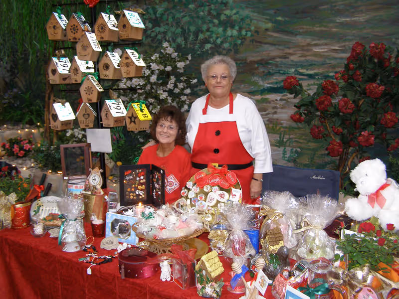 Two elderly women standing behind a table covered with various decorative items and gifts, including wrapped packages, ornaments, and a large white teddy bear. Behind them is a display of small wooden birdhouses and a painted backdrop with greenery and flowers.