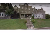 A large, multi-story stone building with a central entrance accessed by a staircase leading up from the street. The building has multiple windows and a covered porch area. There is a grassy lawn in front and some trees on the left side.
