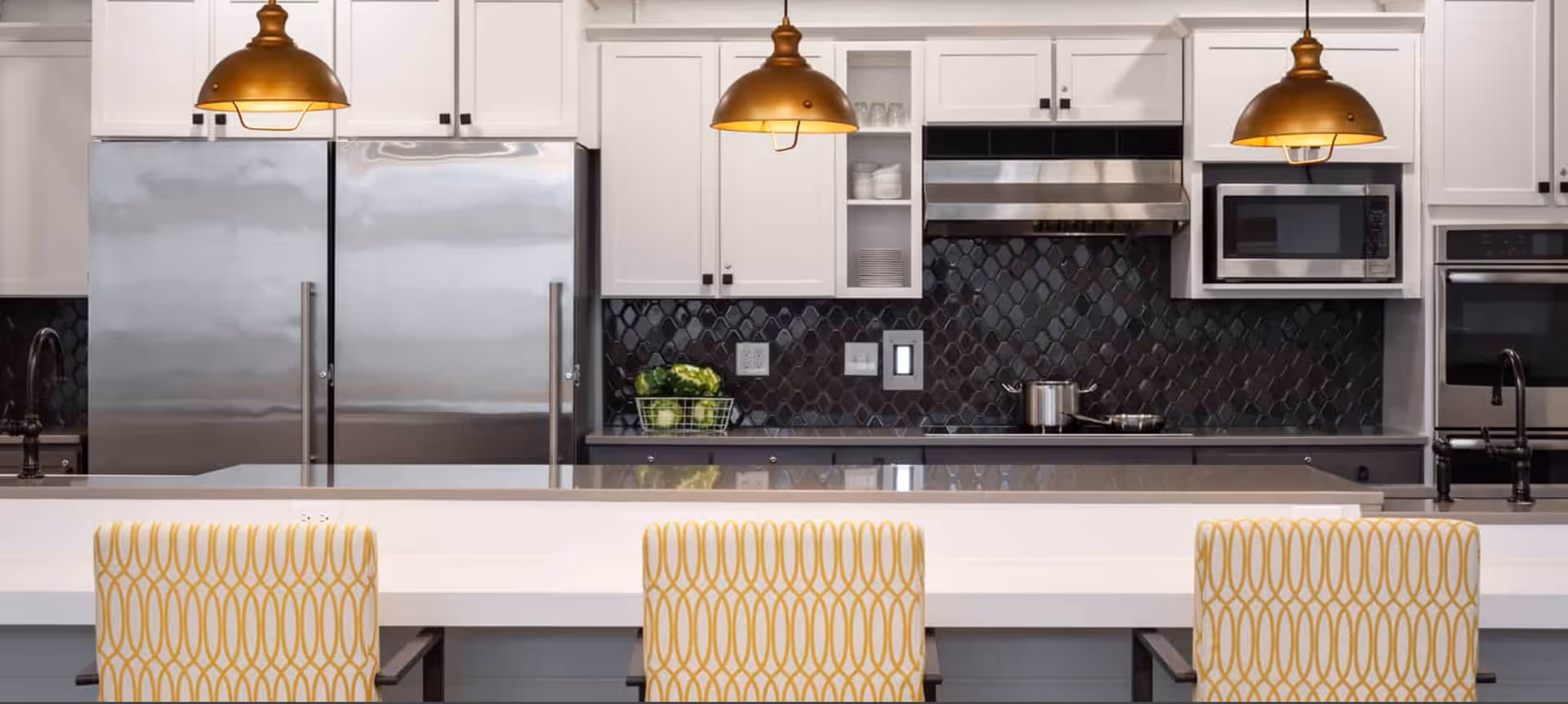 Modern kitchen with white cabinetry, stainless steel refrigerator, microwave, and oven. The backsplash features black tiles with a geometric pattern. Three pendant lights with bronze shades hang over a white countertop island with three yellow patterned chairs.