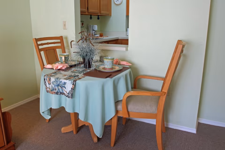 Small dining table set for two with wooden chairs, tablecloth, place settings and a centerpiece beside a kitchen pass-through.