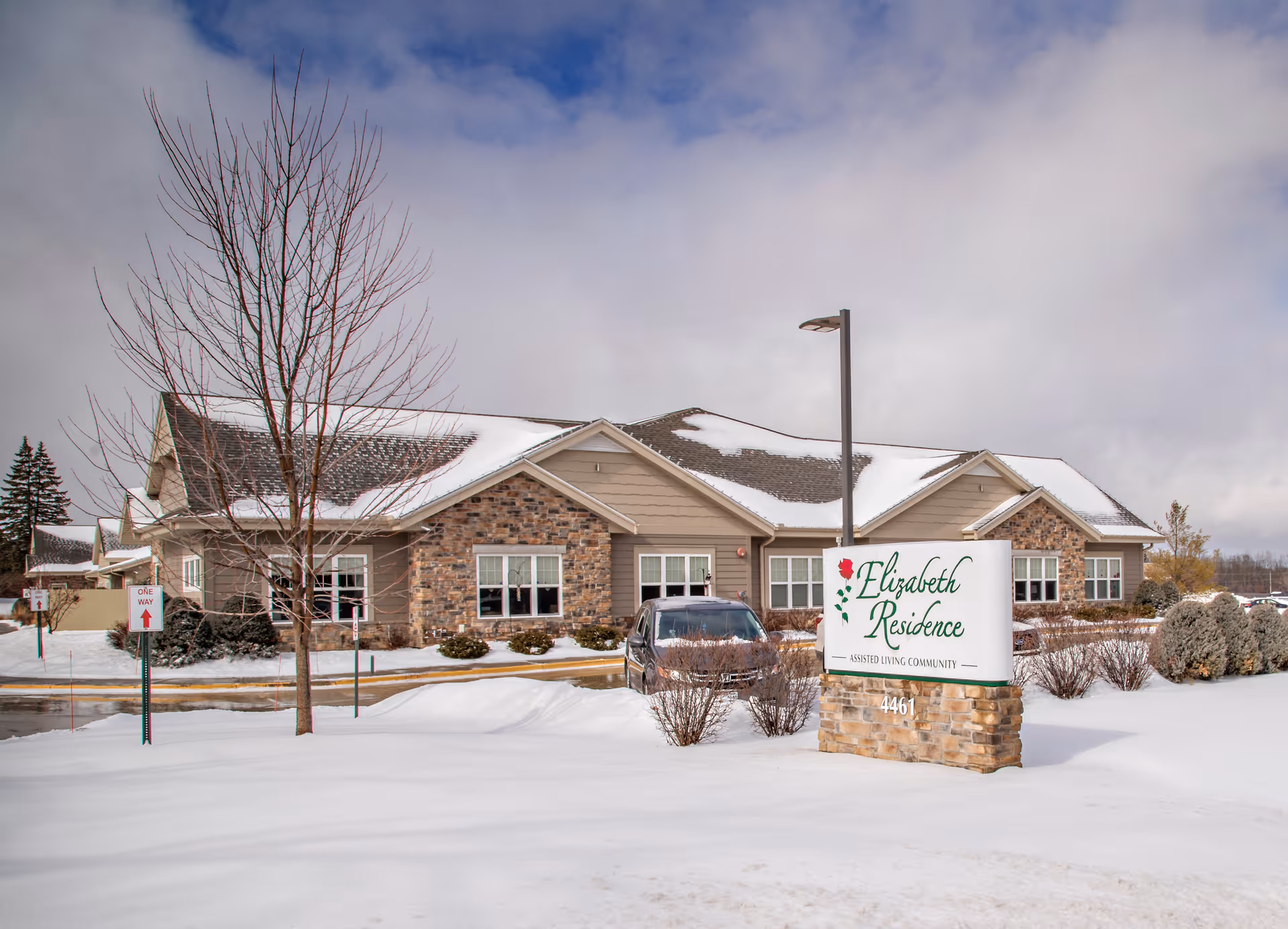 Exterior view of Elizabeth Residence New Berlin, an assisted living community building with a stone and beige siding facade, surrounded by snow-covered ground and a cloudy sky. A sign in front displays the facility name and address 4461.