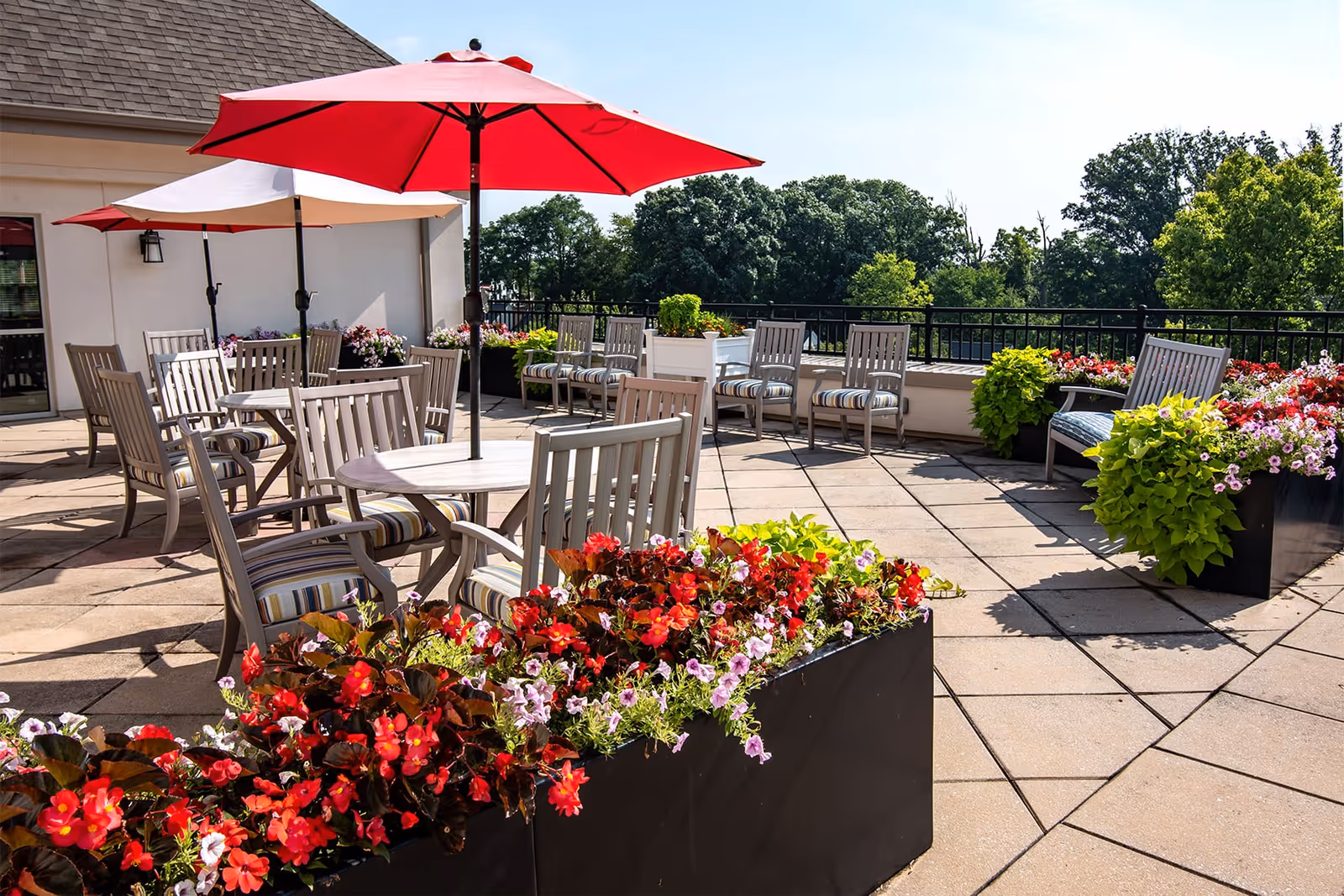 Outdoor patio area with multiple tables and chairs under large red and white umbrellas. The patio is decorated with planters filled with colorful flowers and greenery. Trees and a clear sky are visible in the background.