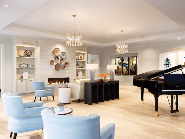 A spacious and elegant living room area with light wood flooring, light blue armchairs, a black grand piano, and a modern fireplace surrounded by decorative plates on the wall. The room features built-in shelves with books and decor, two pendant lights hanging from the ceiling, and framed artwork on the walls.