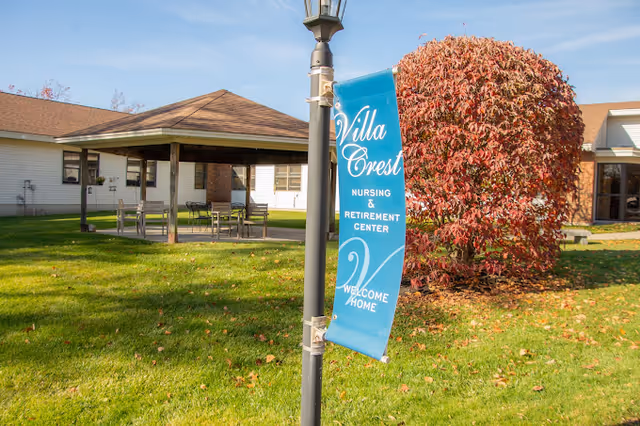 Outdoor view of Villa Crest Nursing and Retirement Center showing a green lawn with a large bush with red leaves, a covered seating area with tables and chairs, and a lamp post with a blue banner that reads 'Villa Crest Nursing & Retirement Center Welcome Home'.