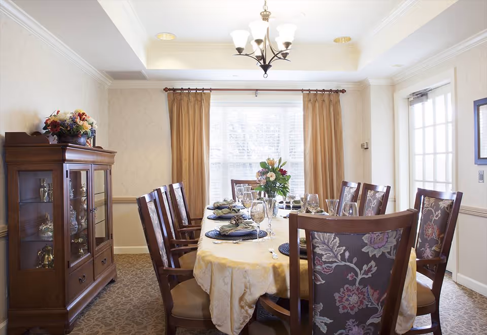Formal dining room with a long set table covered by a tablecloth, wooden chairs with floral upholstery, a china cabinet, chandelier, and curtained window.