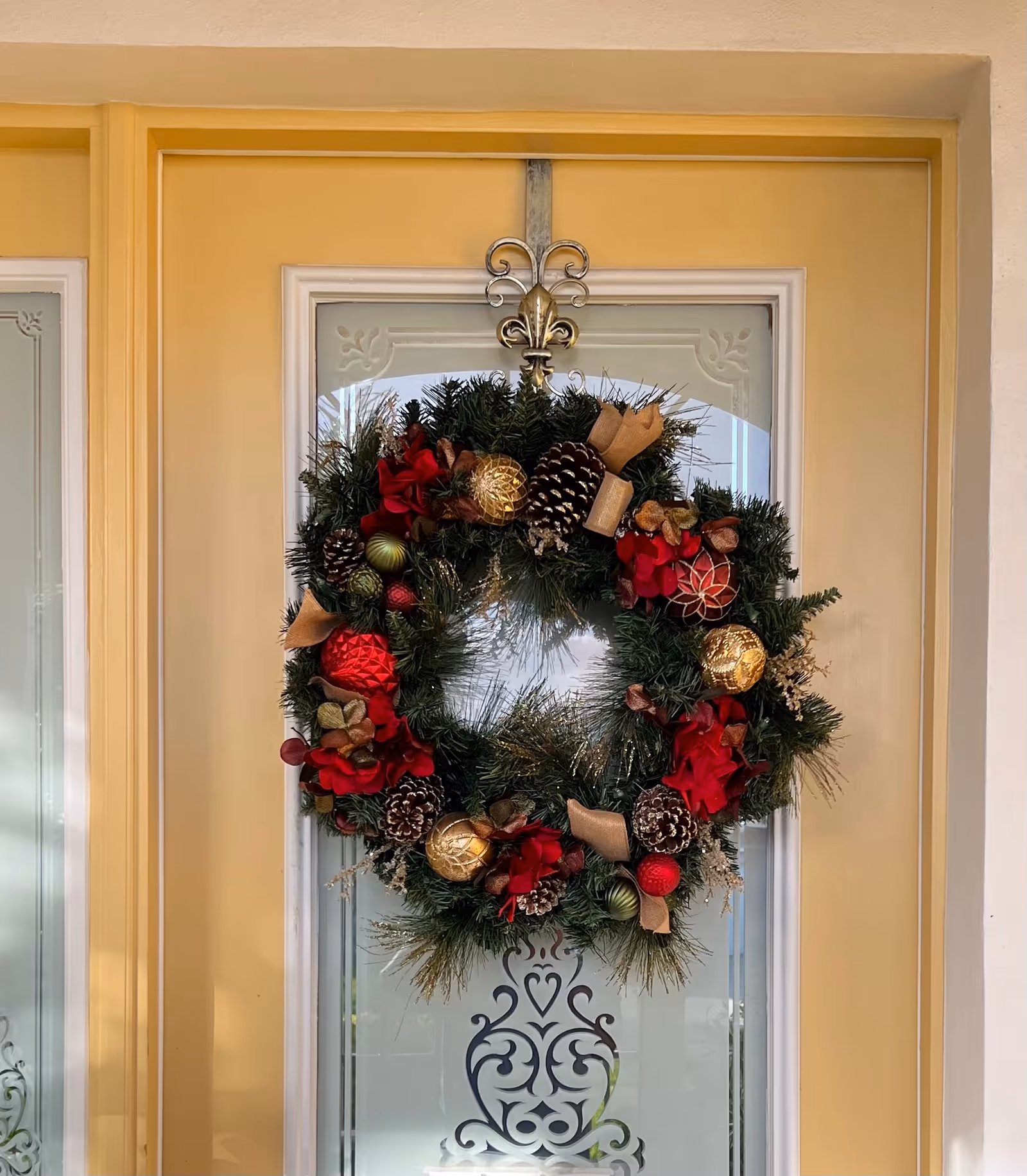 A festive holiday wreath decorated with pine cones, red and gold ornaments, and red flowers hanging on a yellow door with a glass panel featuring an ornate design.