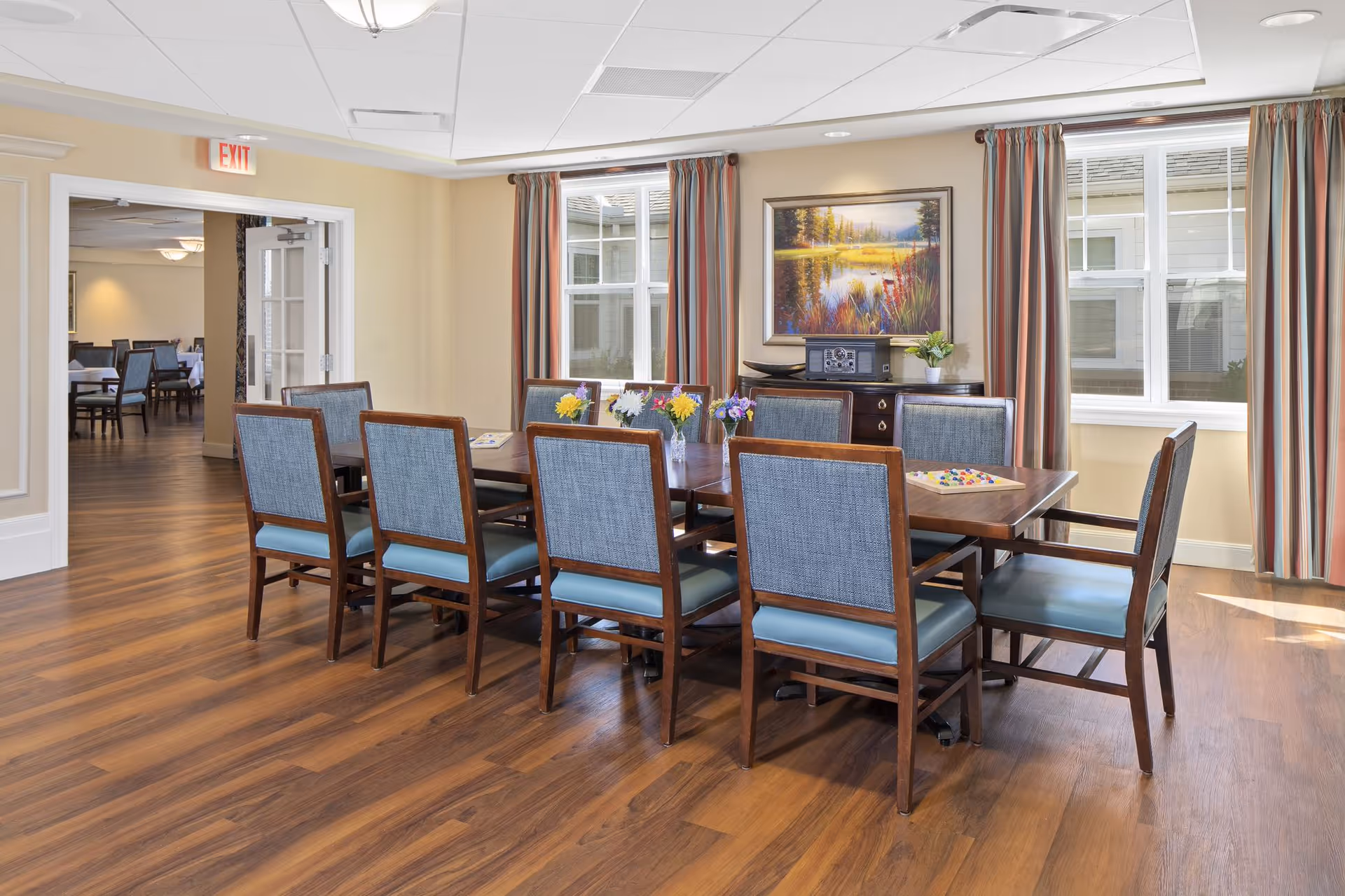 A bright dining room with a wooden table surrounded by eight chairs with blue cushions. The room has wood flooring, large windows with striped curtains, and a painting of a nature scene on the wall. There are small flower arrangements on the table and a sideboard with a radio and a plant underneath the painting.