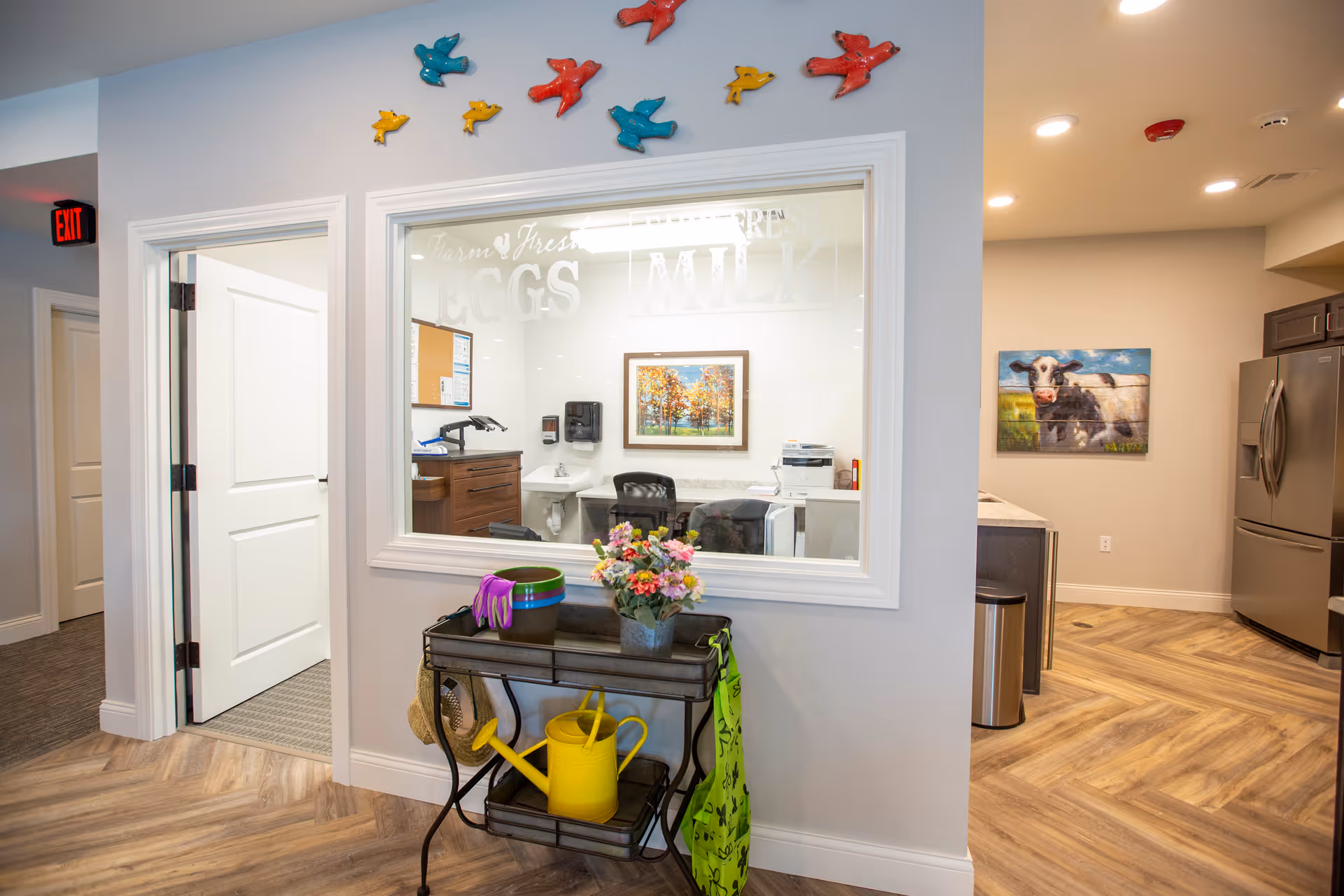 Interior common area with a service window into an office, a small table with flowers and gardening items in front, and a kitchen area with a refrigerator and cow painting.