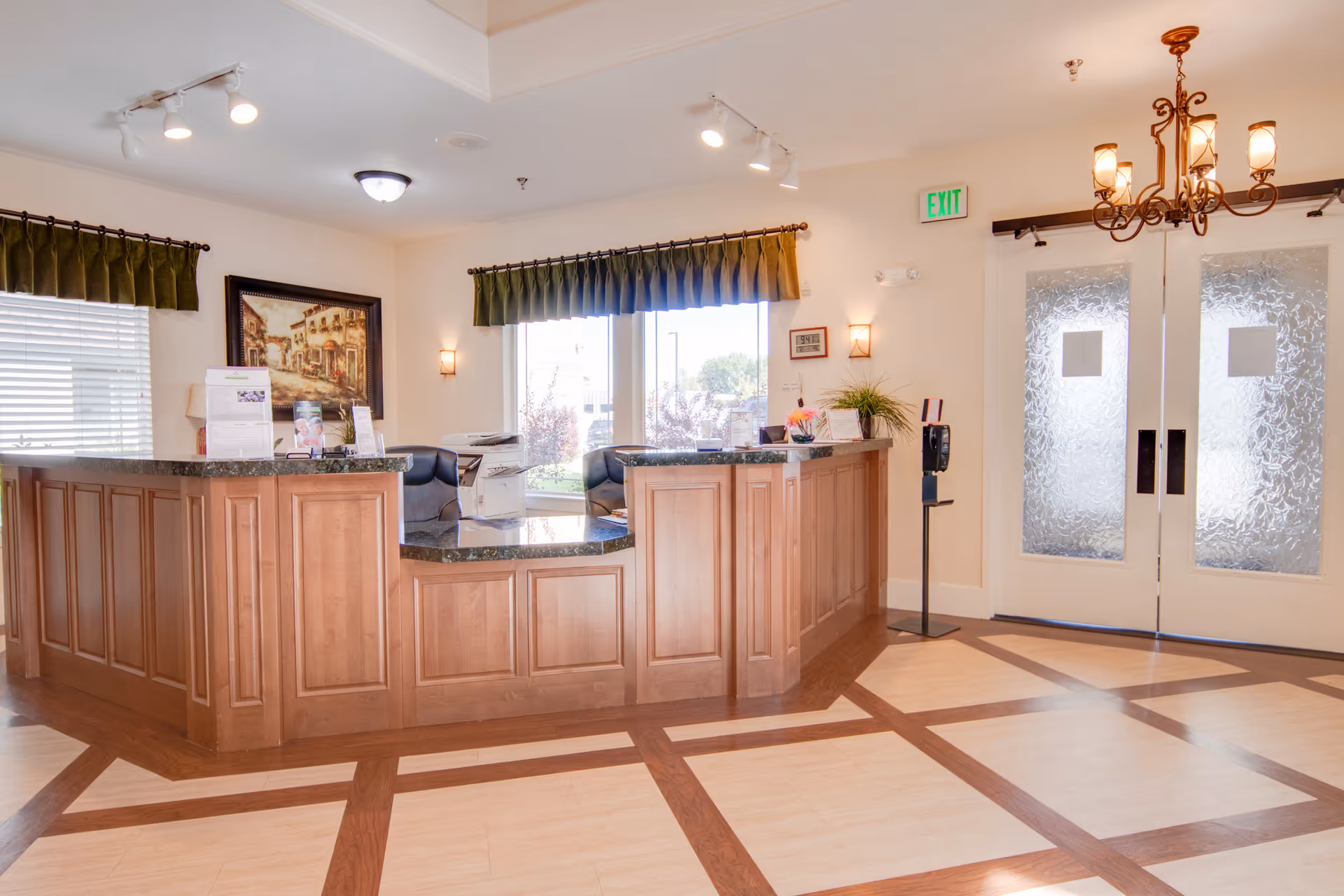 Reception area with a wooden front desk featuring a dark granite countertop, two office chairs behind the desk, a printer, and informational brochures. The room has large windows with green valances, a chandelier, and double frosted glass doors with an exit sign above them. The floor has a light tile pattern with darker wood inlays.