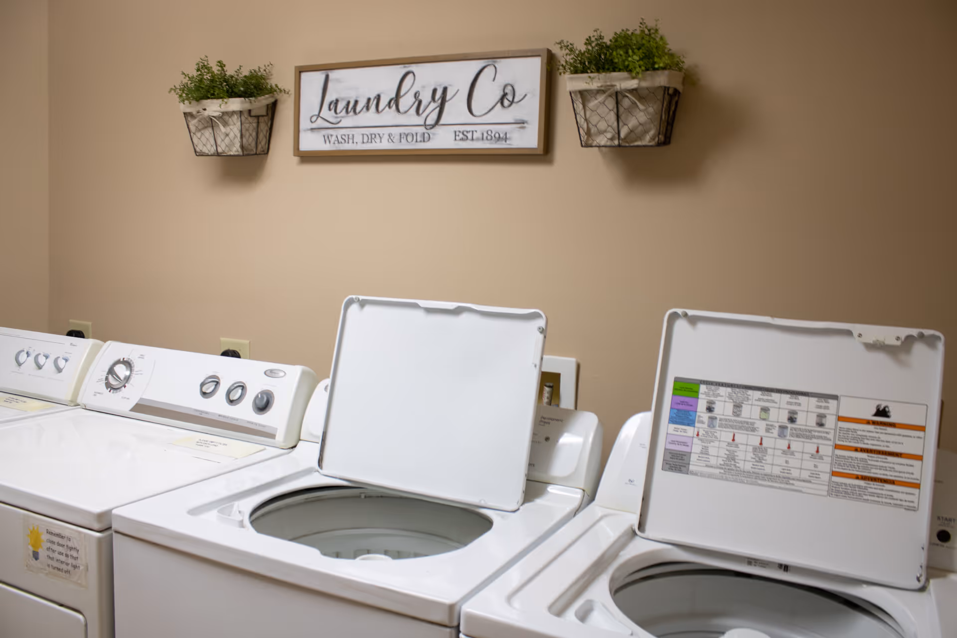 Row of top-loading washing machines in a laundry room with a 'Laundry Co' sign and wall planters.