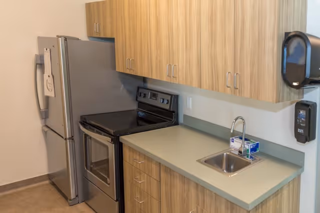 Compact kitchenette with stainless steel refrigerator and stove, laminate countertop with sink, and wood-grain cabinets.
