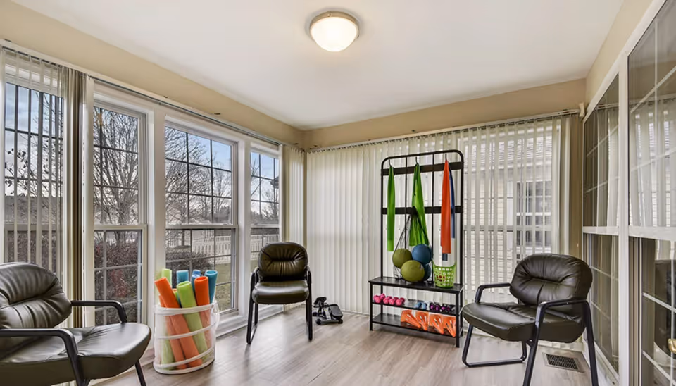 A bright room with large windows and vertical blinds, containing three black cushioned chairs, a basket with colorful foam rollers, and a rack holding exercise balls, resistance bands, and small dumbbells, suggesting a light exercise or therapy space.