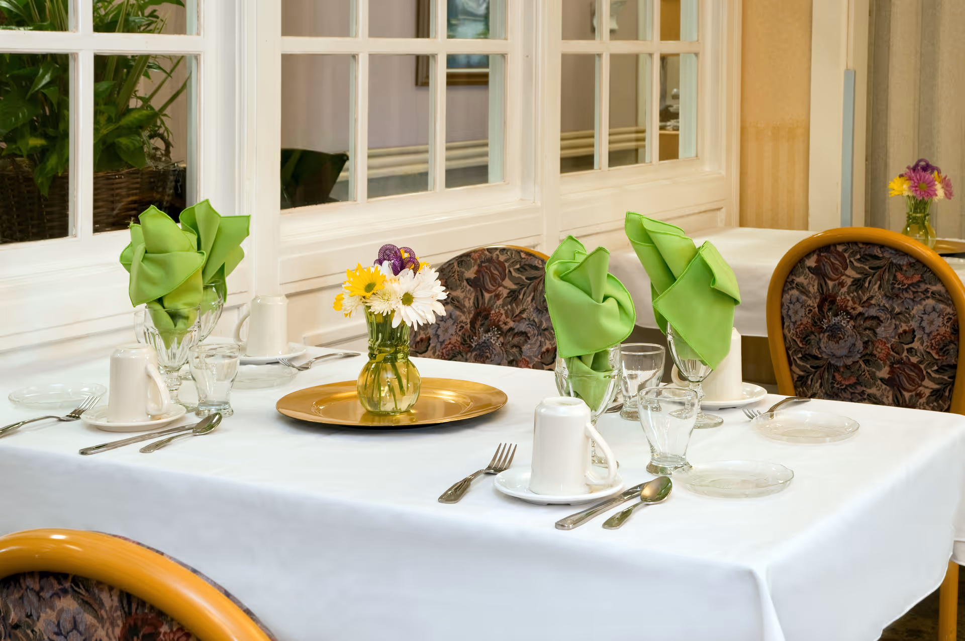 A dining table set with white tablecloth, green folded napkins in glasses, white cups, plates, silverware, and a small vase with yellow and white flowers. The table is surrounded by chairs with floral upholstery, and there is a window with white framing in the background.