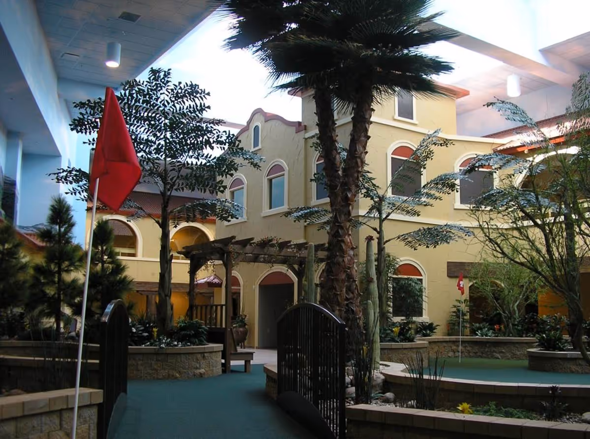 Indoor courtyard area with a putting green, palm trees, other plants, a small arched bridge, and a building with arched windows and a yellow exterior. There are red flags marking holes on the putting green.