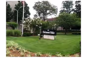 Outdoor view of a green lawn with trees and shrubs surrounding a sign that reads 'Clairmont Oaks'. A building and flagpole are visible in the background.