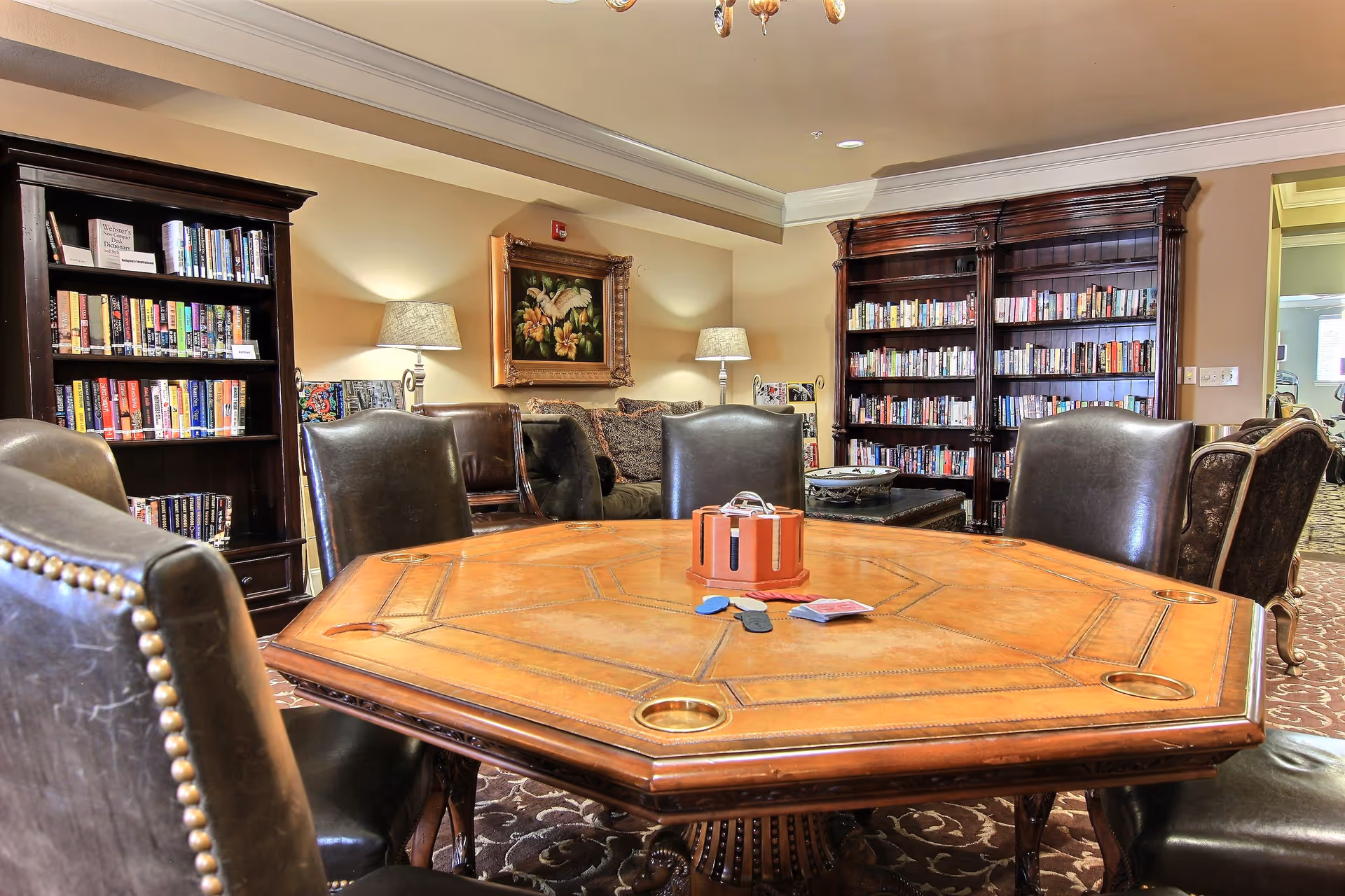 A cozy interior room featuring a large wooden octagonal table surrounded by six leather chairs. In the background, there are two tall dark wood bookshelves filled with books, a sofa with cushions, two table lamps, and a framed floral painting on the wall. The room has warm lighting and a patterned carpet.