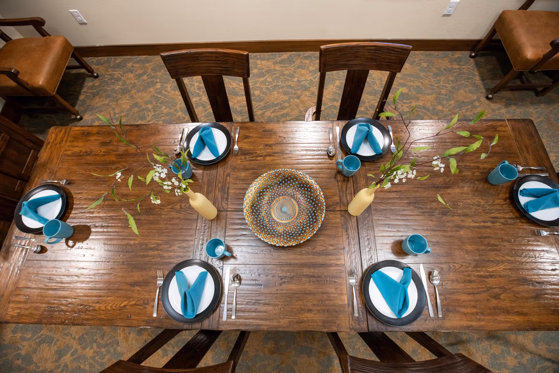 Overhead view of a wooden dining table set for six with teal napkins and mugs, vases with branches, and a decorative centerpiece.
