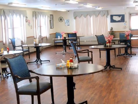 Bright dining room with round tables, chairs, and windows with drapes in a senior living facility.