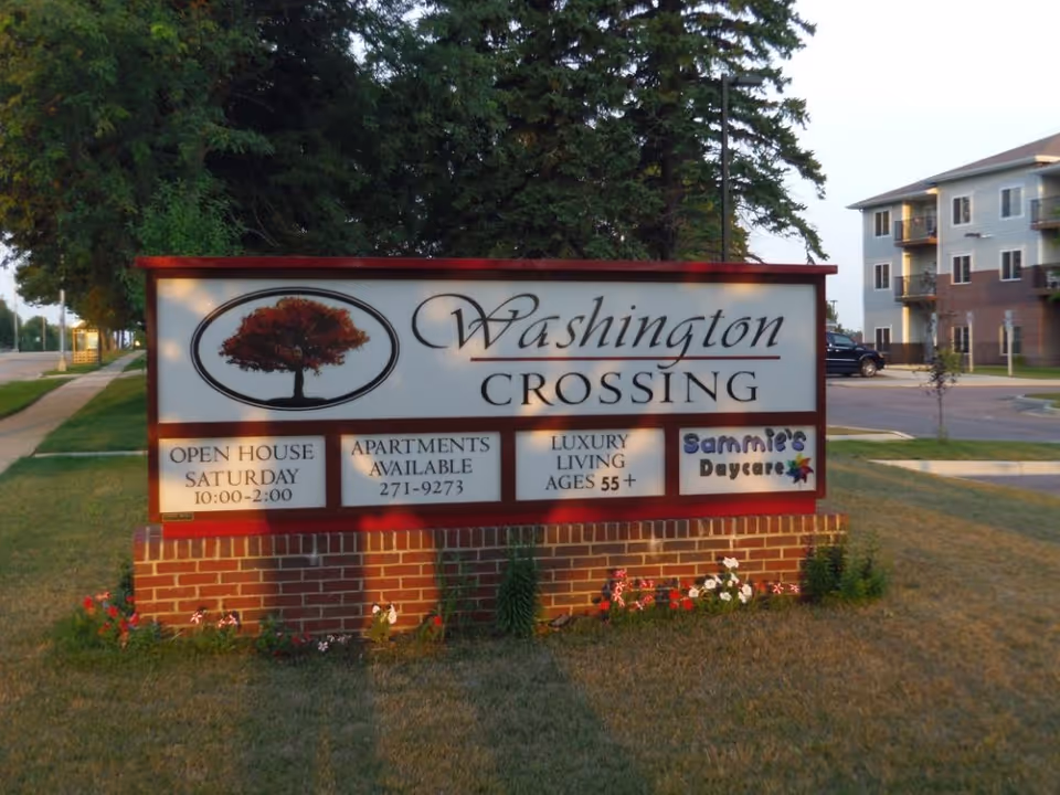 Outdoor sign for Washington Crossing featuring a tree logo, advertising an open house on Saturday from 10:00 to 2:00, apartments available with phone number 271-9273, luxury living for ages 55+, and Sammie's Daycare. The sign is set on a brick base with grass and flowers around it, with trees and a building visible in the background.