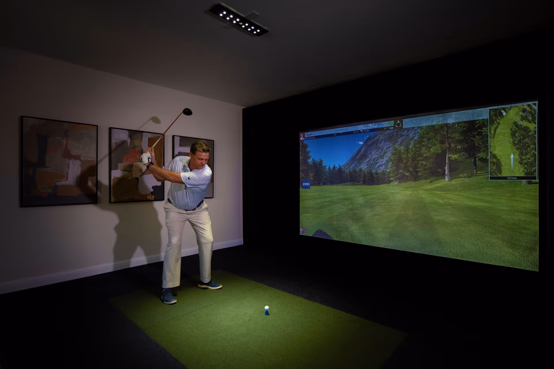 A man is playing indoor golf using a golf simulator. He is in a room with a green mat on the floor, preparing to swing a golf club towards a golf ball on a tee. A large screen in front of him displays a virtual golf course with trees and mountains.