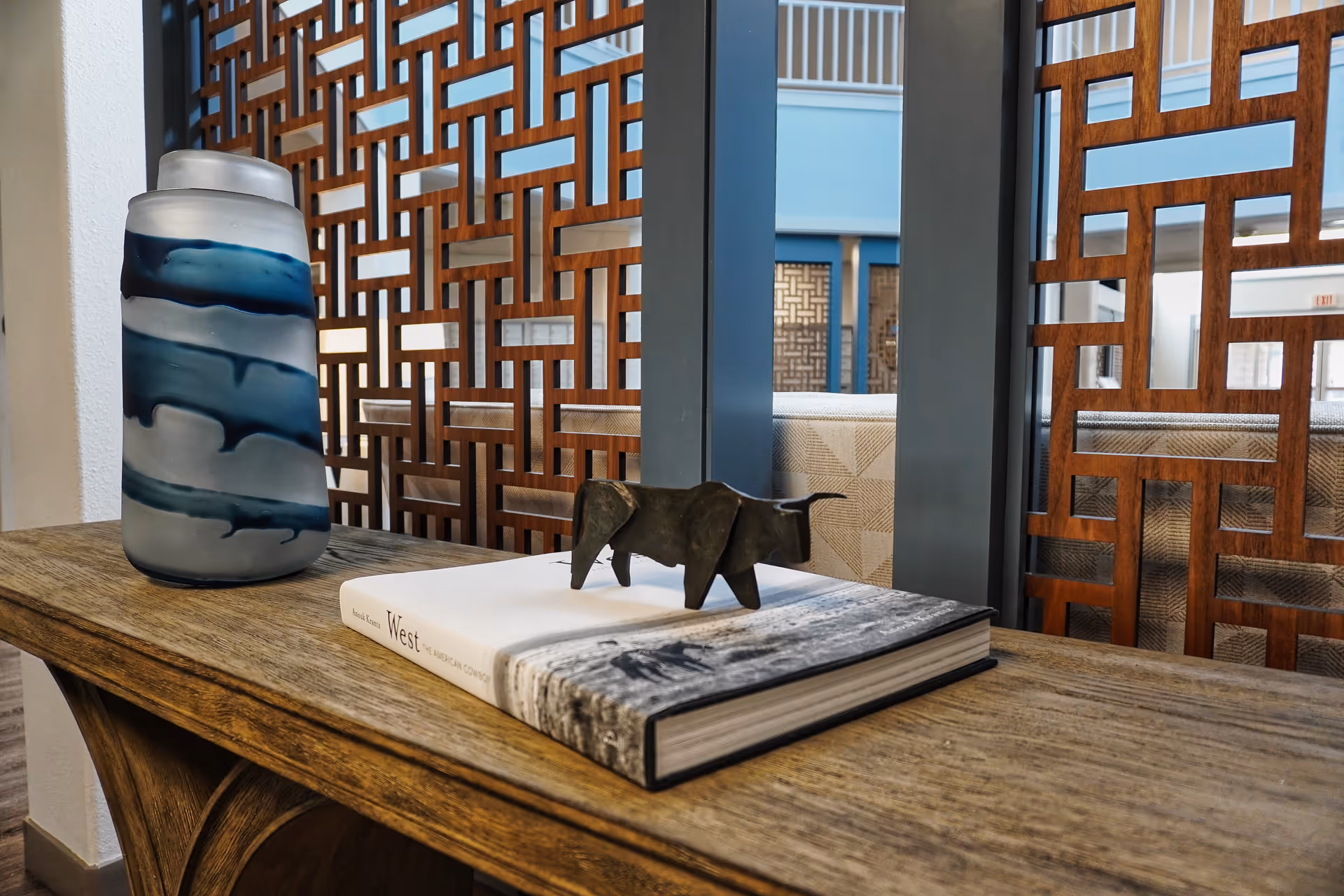 A decorative wooden table with a blue and white glass vase, a book titled 'West: The American Cowboy', and a small metal bull figurine. Behind the table is a wooden lattice room divider with geometric patterns, and a glimpse of a modern interior space with blue and white walls.