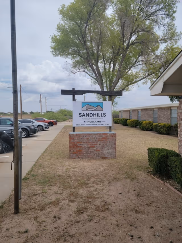 Outdoor view of Sandhills Nursing and Rehabilitation facility sign mounted on a brick base with a tree and building in the background, and cars parked along a driveway to the left.