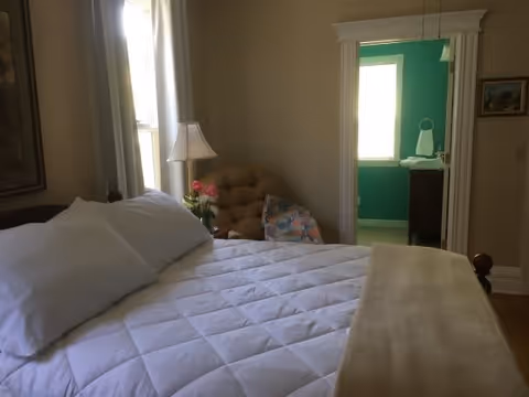 A cozy bedroom with a neatly made bed featuring white pillows and a white quilt. Next to the bed is a small table with a lamp and a vase of flowers. In the corner, there is a cushioned chair with a colorful pillow. Through an open doorway, a bathroom with teal walls and a window is visible.