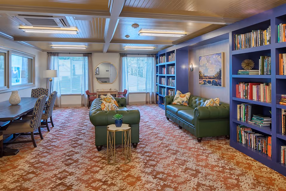 A cozy living room with two green leather sofas adorned with floral pillows, a small round side table with a potted plant, blue built-in bookshelves filled with books, a painting on the wall, a long wooden table with patterned chairs on the left, and two large windows with sheer curtains letting in natural light.
