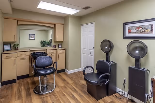Interior view of a salon area in a senior living facility featuring two black salon chairs, two hair dryer stations, wooden cabinetry with a large mirror, and light green walls with a framed picture.