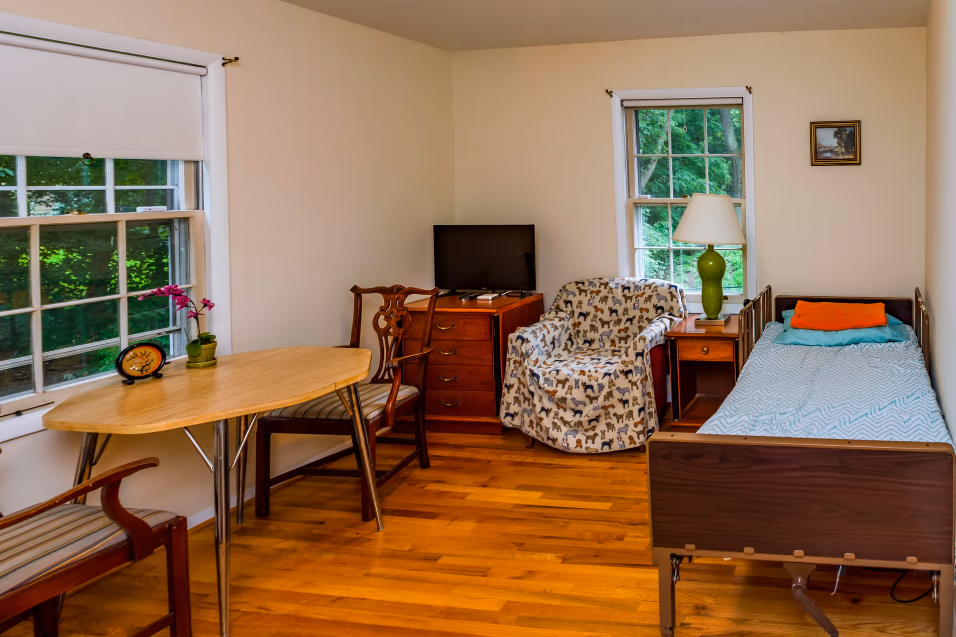 A small, cozy room with a single bed covered with a blue blanket and orange pillow, a wooden nightstand with a green lamp, a patterned armchair, a wooden dresser with a TV on top, and a wooden table with two chairs near two windows showing green trees outside.
