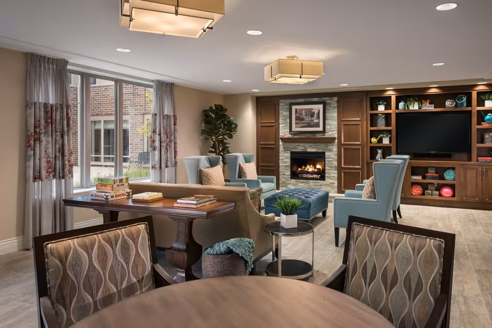 Spacious furnished communal living room with multiple chairs, a fireplace, built-in shelving with a TV, and a table by a large window.