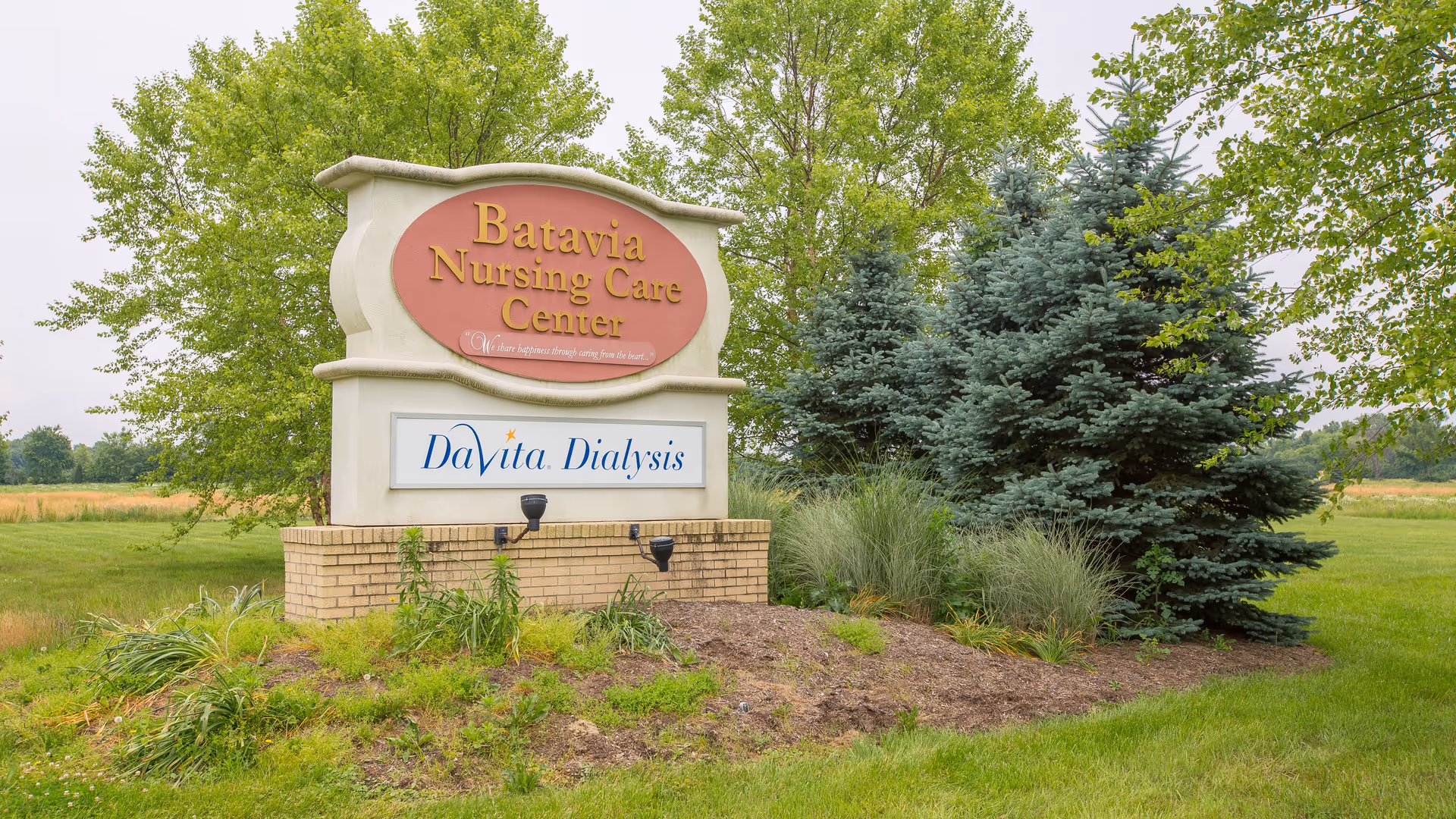 Outdoor view of a sign for Batavia Nursing Care Center and DaVita Dialysis surrounded by greenery, trees, and grass.