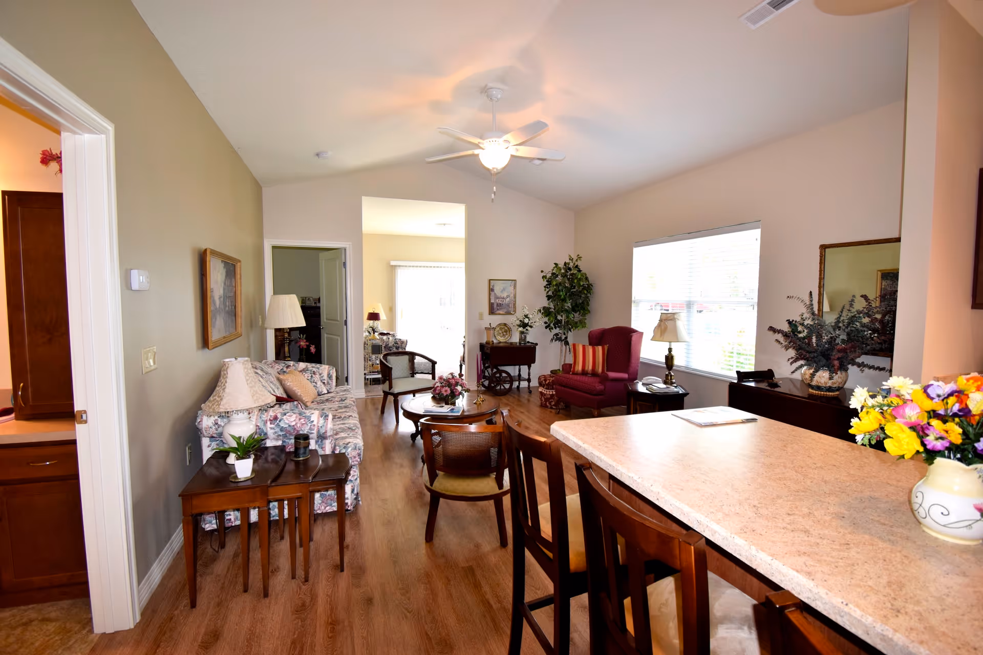 Interior view of a senior living facility's common area with a floral-patterned sofa, wooden chairs, a round coffee table with flowers, a red armchair with a striped pillow, a side table with a lamp, and a countertop with bar stools and a vase of flowers. The room has wood flooring, a ceiling fan with light, and large windows letting in natural light.