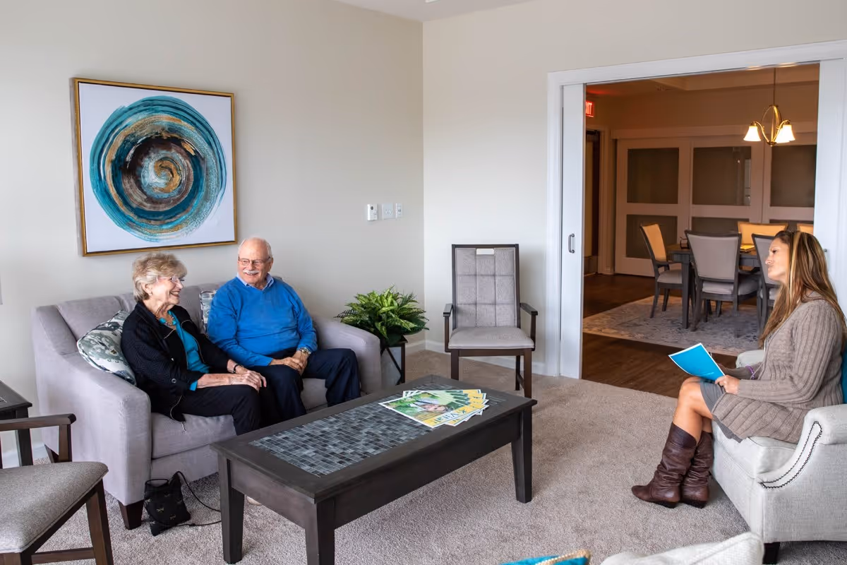 An older couple sits on a couch talking with a younger woman seated across from them in a furnished living room area.