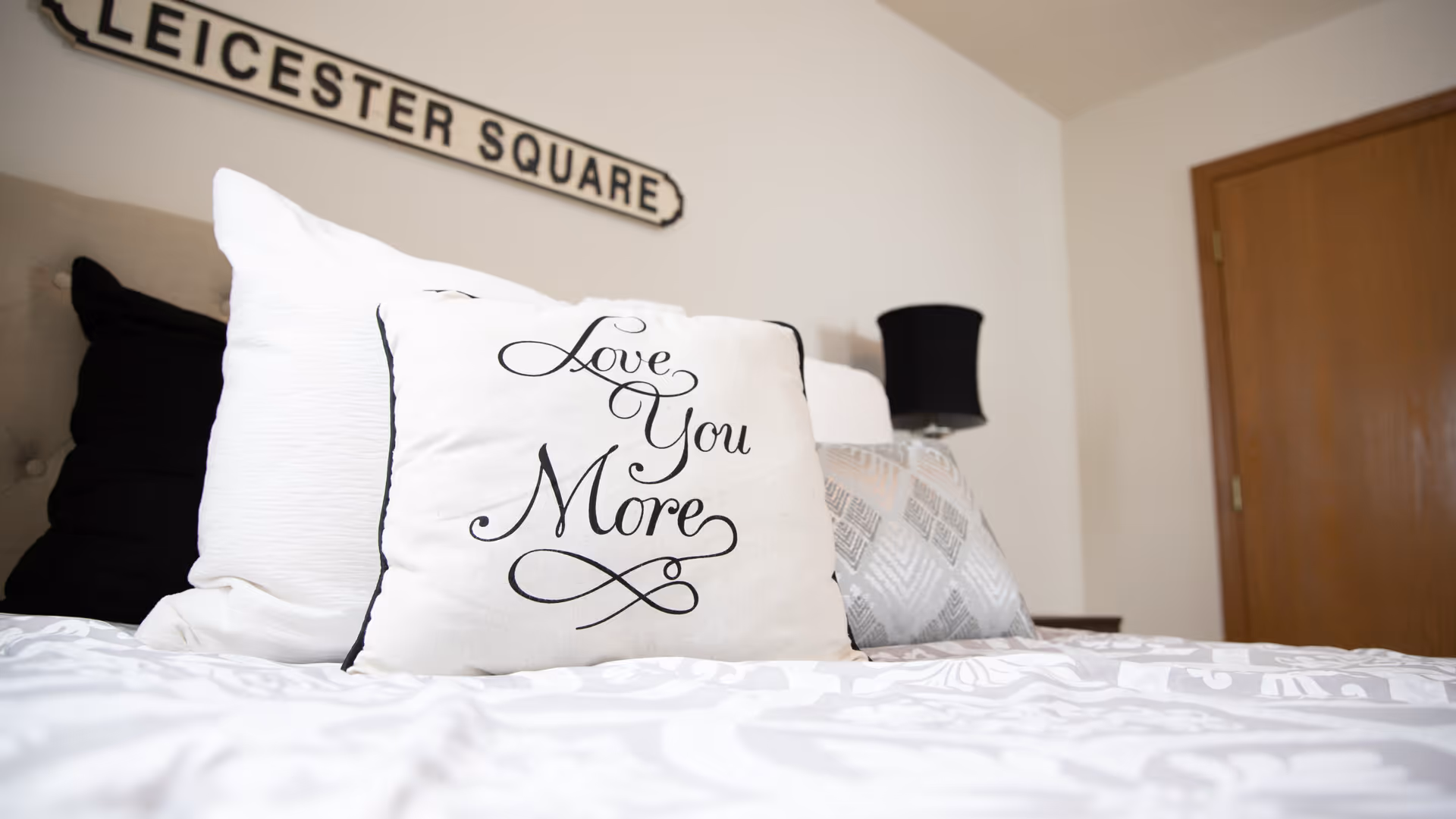 Close-up view of a bed with decorative pillows, including one with the text 'Love You More'. A black lamp and a wooden door are visible in the background, along with a wall sign that reads 'LEICESTER SQUARE'.