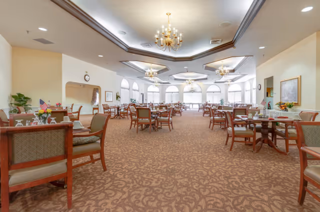 Spacious dining room in an assisted living facility with multiple wooden tables and chairs arranged neatly on a patterned carpet. The room features large arched windows allowing natural light, elegant chandeliers hanging from a coffered ceiling, and decorative plants and framed artwork on the walls.