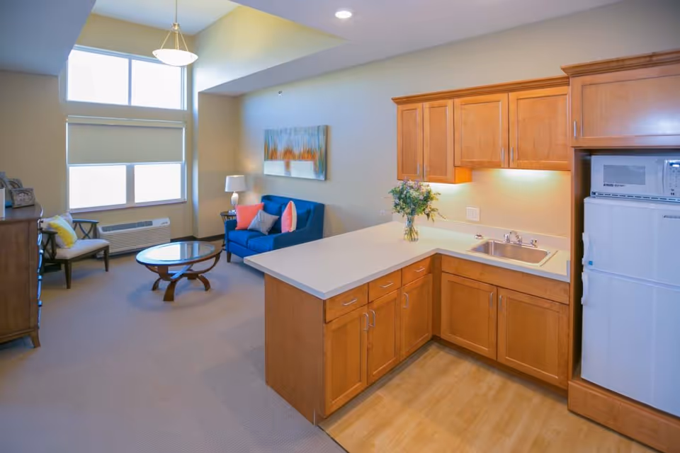 Bright studio-style living area with a small kitchenette, blue sofa, coffee table, and large windows.