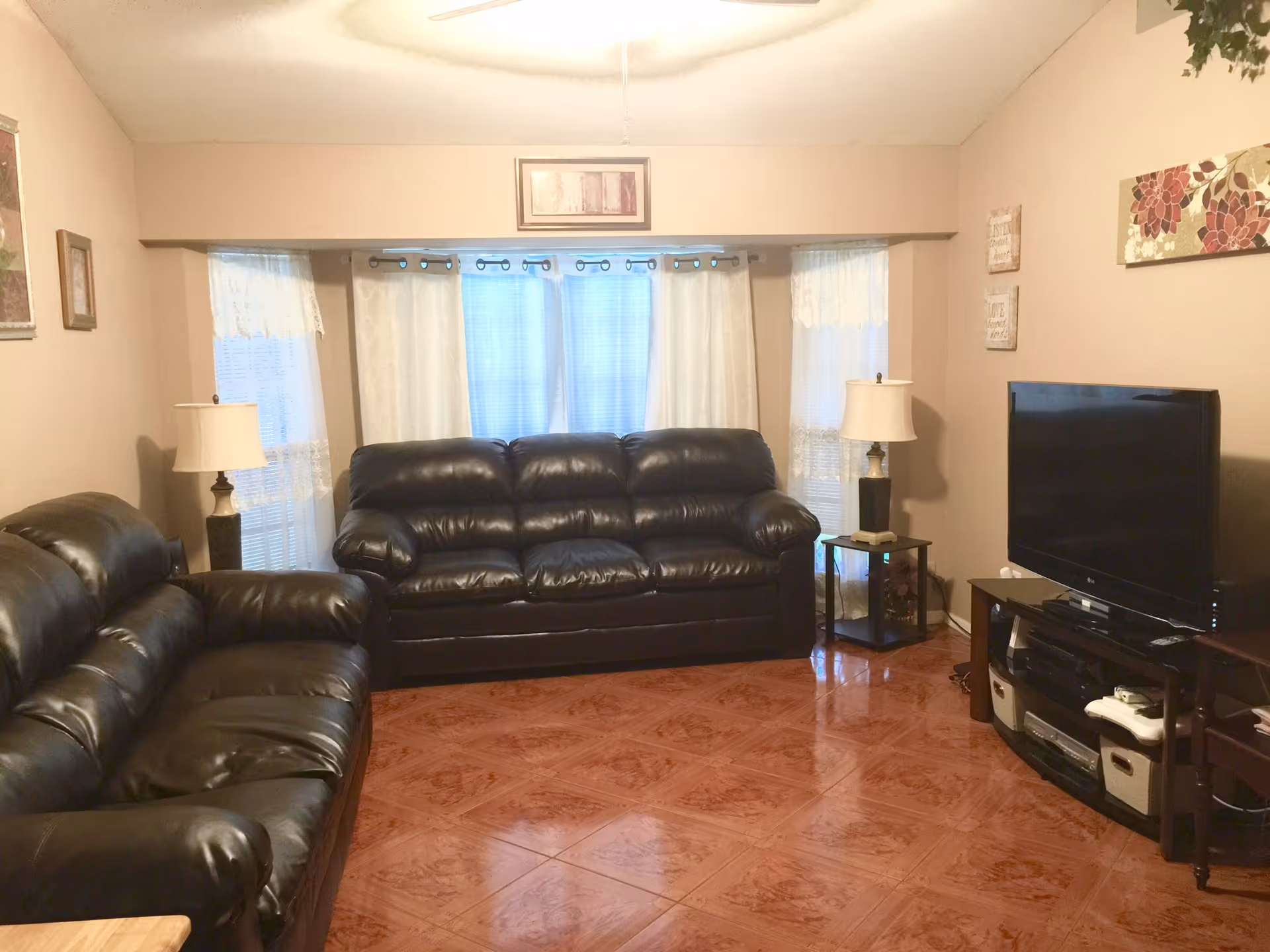 A living room with two black leather sofas facing each other, a flat-screen TV on a stand with shelves holding electronic devices and storage bins, two side tables each with a lamp, and windows with sheer white curtains letting in natural light.