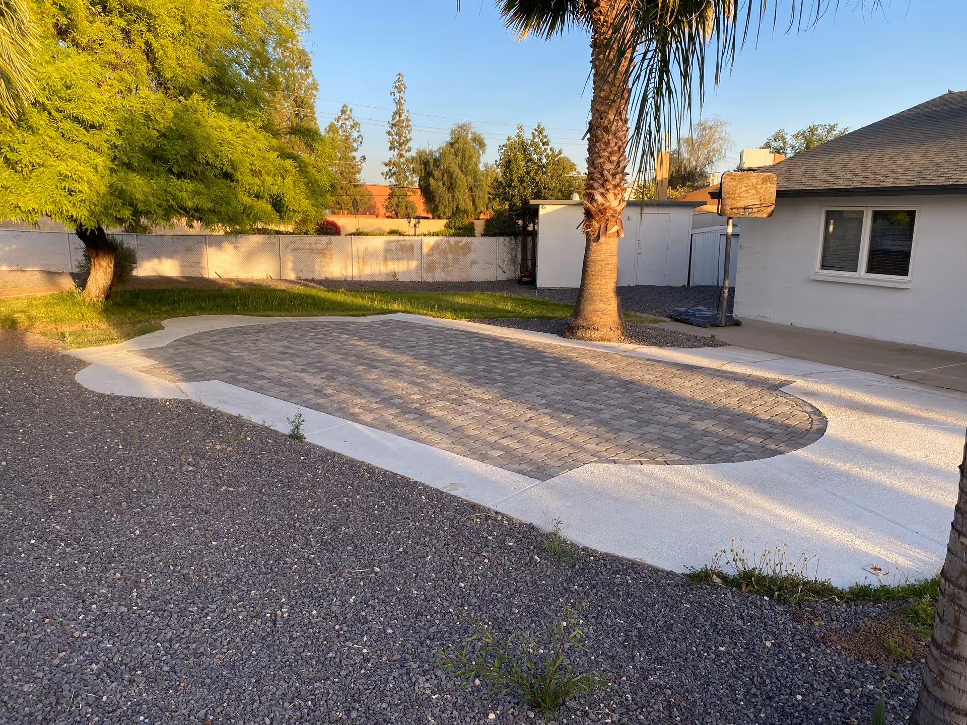 Stone-paved driveway area with a palm tree beside a white single-story house and surrounding yard under a clear sky.