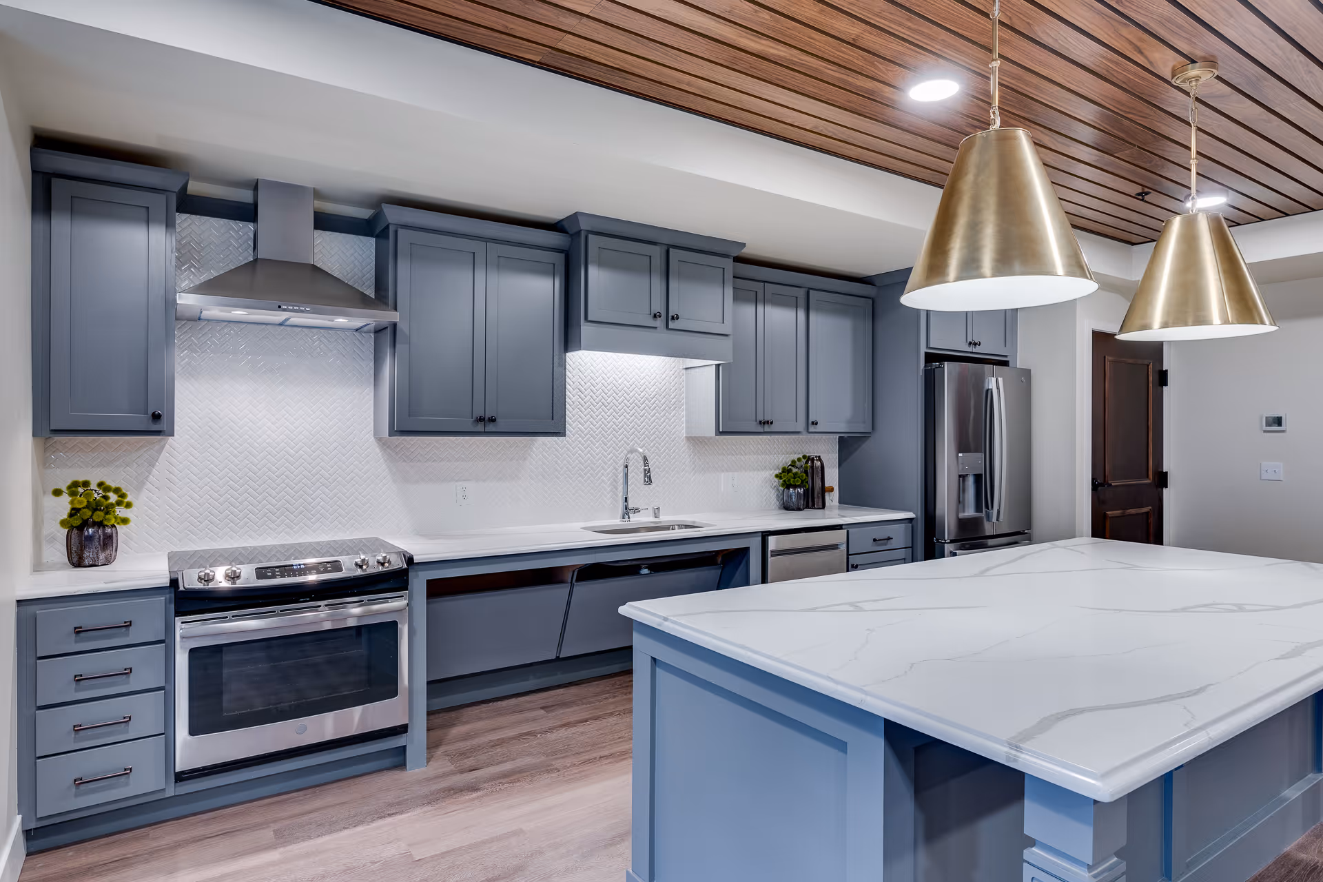 Modern kitchen with blue-gray cabinets, a stainless steel oven and refrigerator, a white marble countertop island, and two large brass pendant lights hanging from a wooden ceiling.
