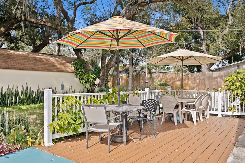 Outdoor patio area with wooden deck featuring two tables with striped umbrellas and various chairs around them. The area is surrounded by a white fence, greenery, and trees under a clear blue sky.