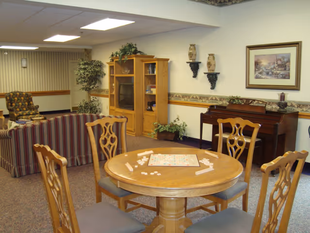 A cozy living room area in a senior living facility featuring a round wooden table with four chairs and a Scrabble game set up on the table. In the background, there is a wooden entertainment center with a TV, a piano against the wall, a framed painting, decorative vases on wall shelves, and some potted plants. The room has carpeted floors and neutral-colored walls with a decorative border near the ceiling.