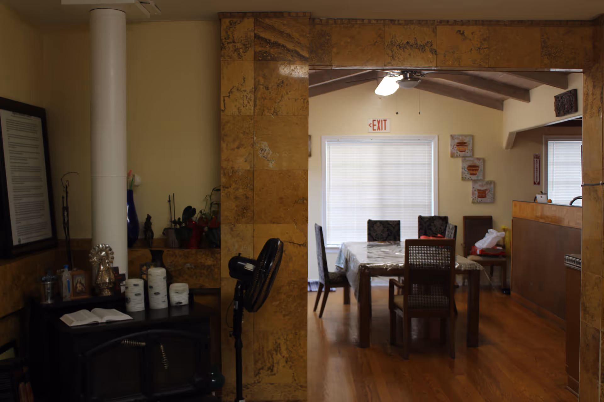 Interior view of a dining area with a wooden table covered by a tablecloth and surrounded by chairs. The room has a window with closed blinds and an exit sign above it. To the left, there is a small black stove with various items on top, including rolls of toilet paper, a fan, and decorative objects. The walls and ceiling beams are beige and brown, and the floor is wooden.