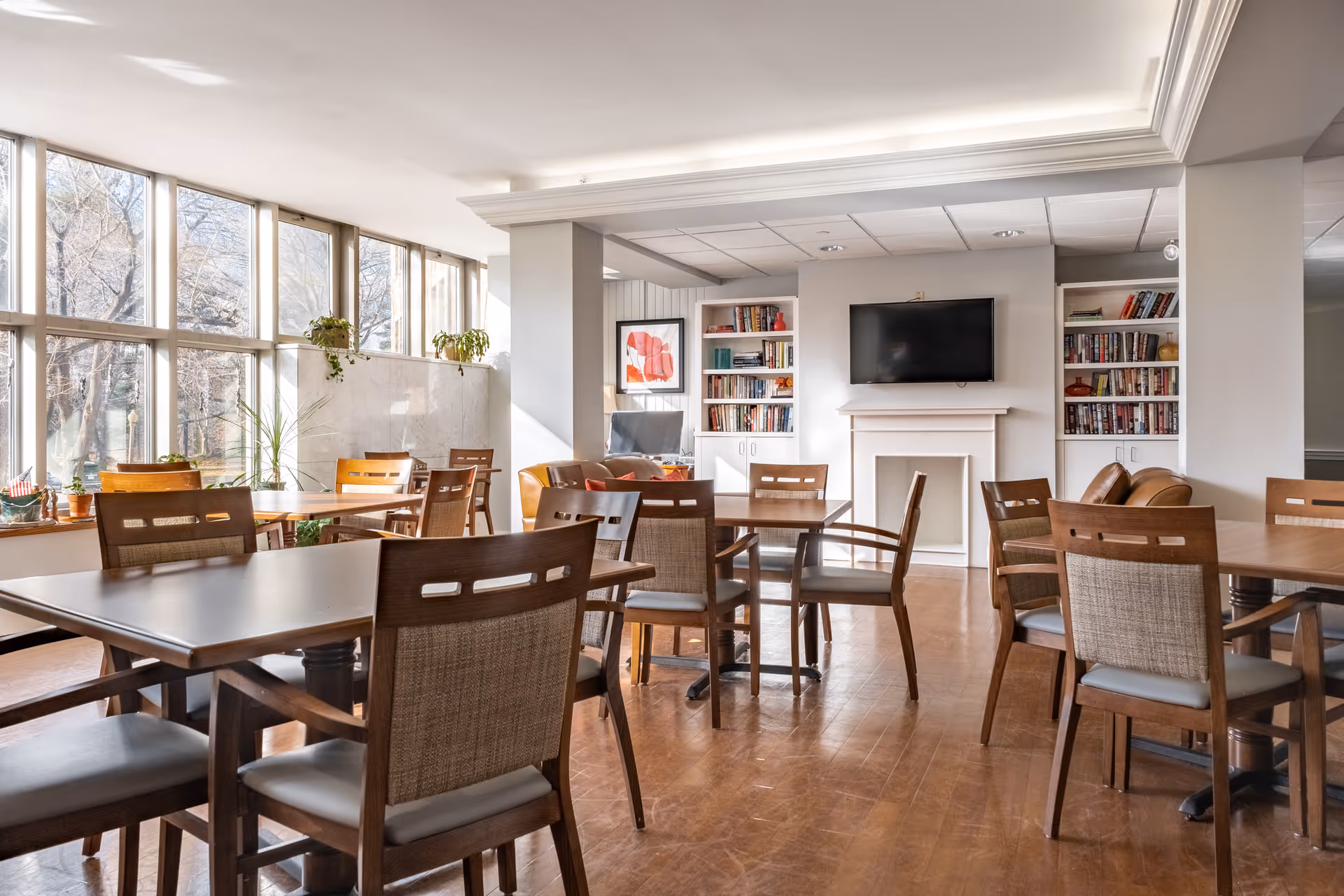 Bright communal dining and lounge area with wooden tables and chairs, large windows, bookshelves, and a wall-mounted TV.