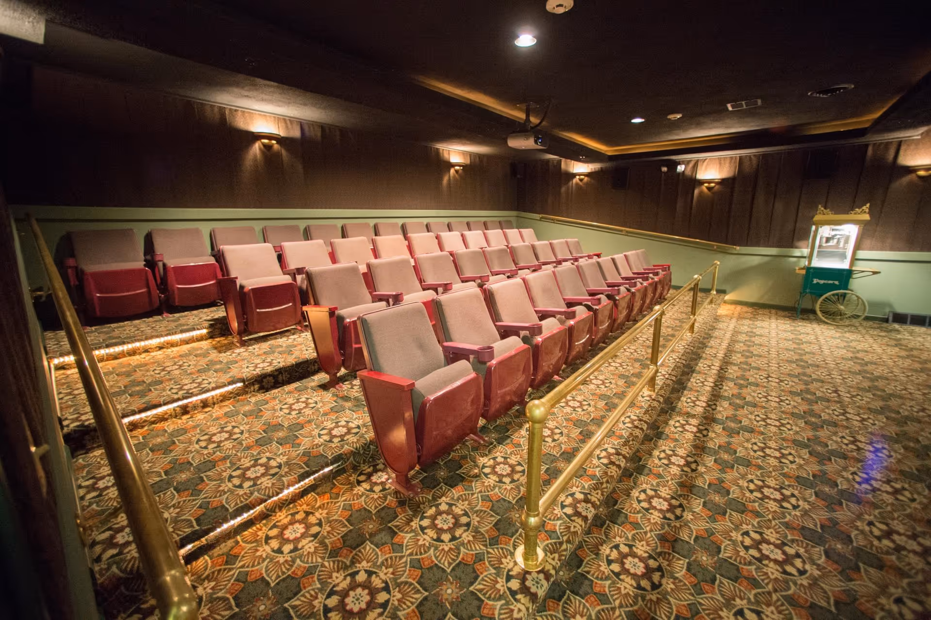 Interior view of a small theater room with rows of red and beige cushioned seats arranged in tiered rows. The room has patterned carpet flooring, dim wall lighting, and a popcorn machine on the right side near the wall.
