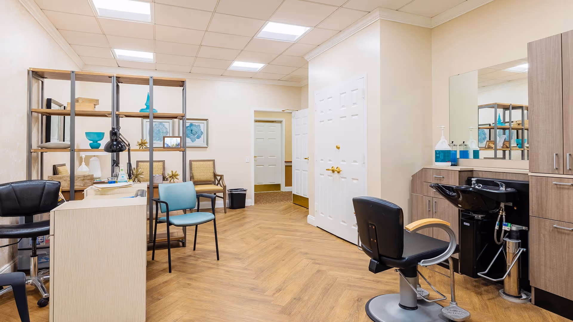 A bright and clean salon area inside a senior living facility with wooden flooring, a black salon chair in front of a hair washing station, a blue chair, and a black rolling chair. There is a shelving unit with decorative items and two wooden chairs against the wall. The room has white walls, a large mirror above the hair washing station, and a white double door in the background.