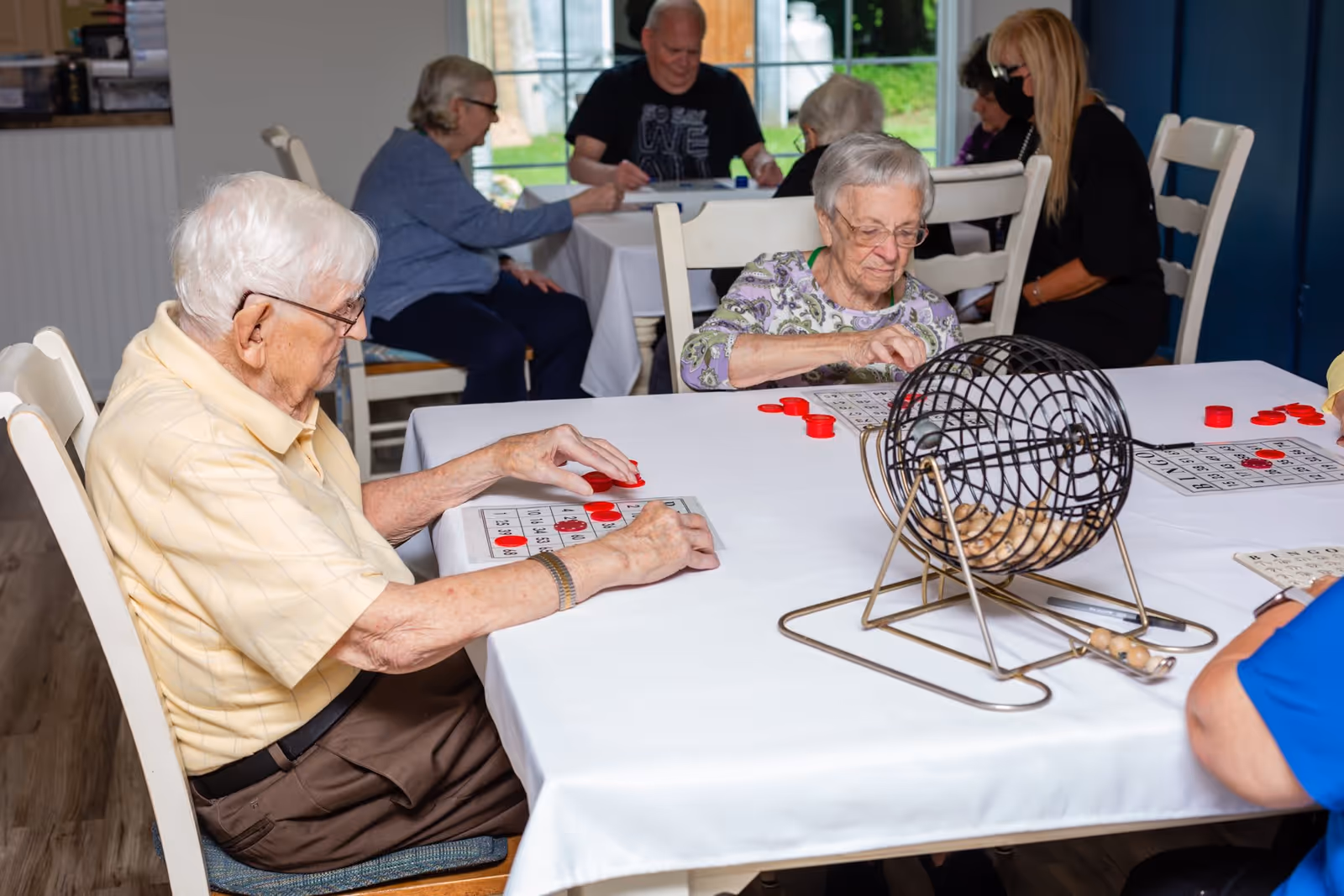 A group of elderly people sitting around tables playing bingo in a well-lit room with large windows. They are placing red markers on their bingo cards, and a bingo cage with numbered balls is on the table.