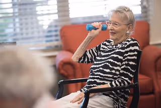 An elderly woman wearing glasses and a black and white striped shirt is sitting in a chair and lifting a small blue dumbbell in a bright room with large windows and a red armchair in the background.