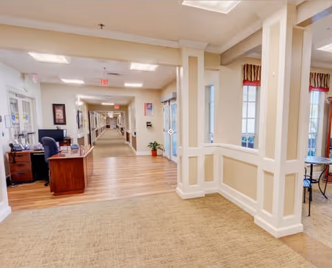 Interior view of a senior living facility hallway with a reception desk on the left, beige carpet and wood flooring, cream-colored walls with white trim, and a seating area with windows on the right.